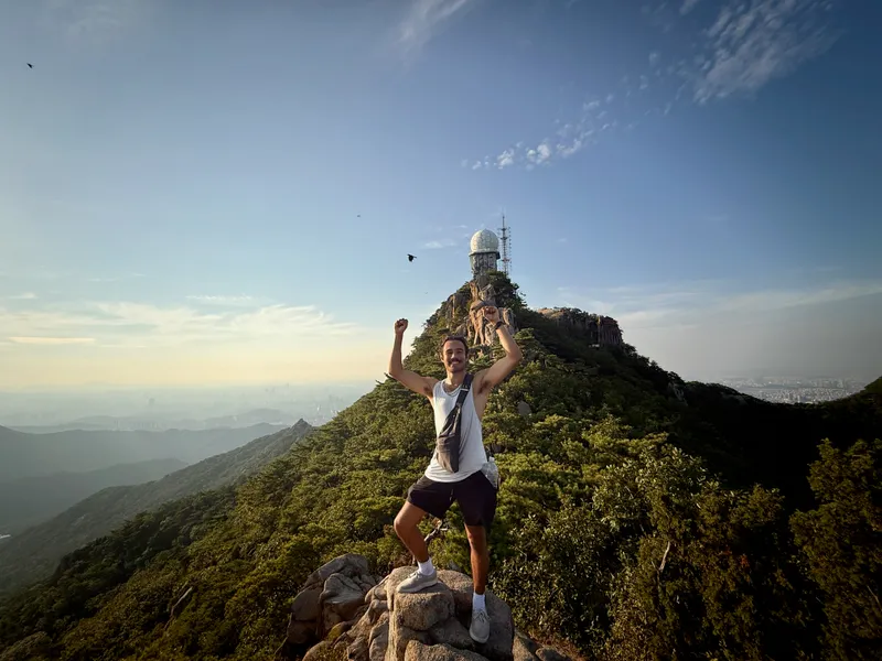 Karl with fists raised on a mountain summit near Seoul, radar dome visible in the background
