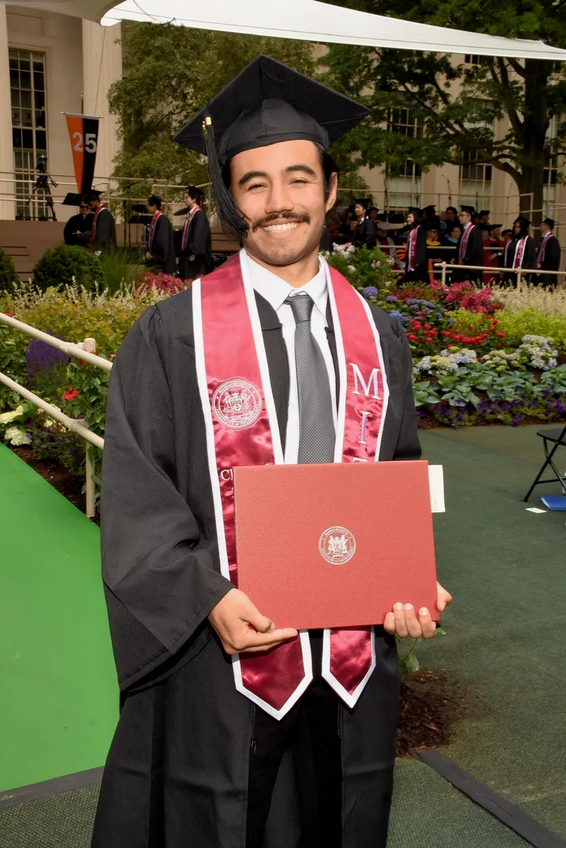 Karl in MIT cap and gown holding his diploma case in Killian Court