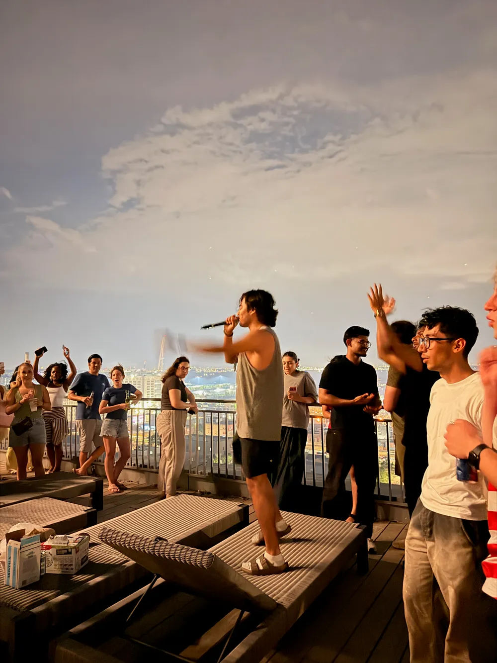 Karl giving an announcement into a microphone at a rooftop party he hosted, crowd cheering around him, NYC skyline at dusk