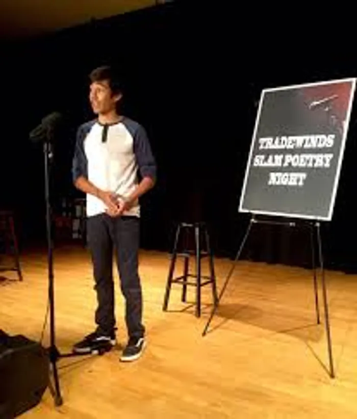 A younger Karl standing alone on stage at a microphone, Tradewinds Slam Poetry Night sign beside him